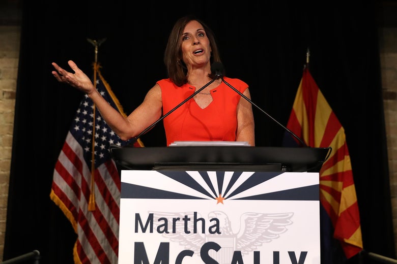 Martha McSally, in a red dress, gestures upward while speaking at a podium.
